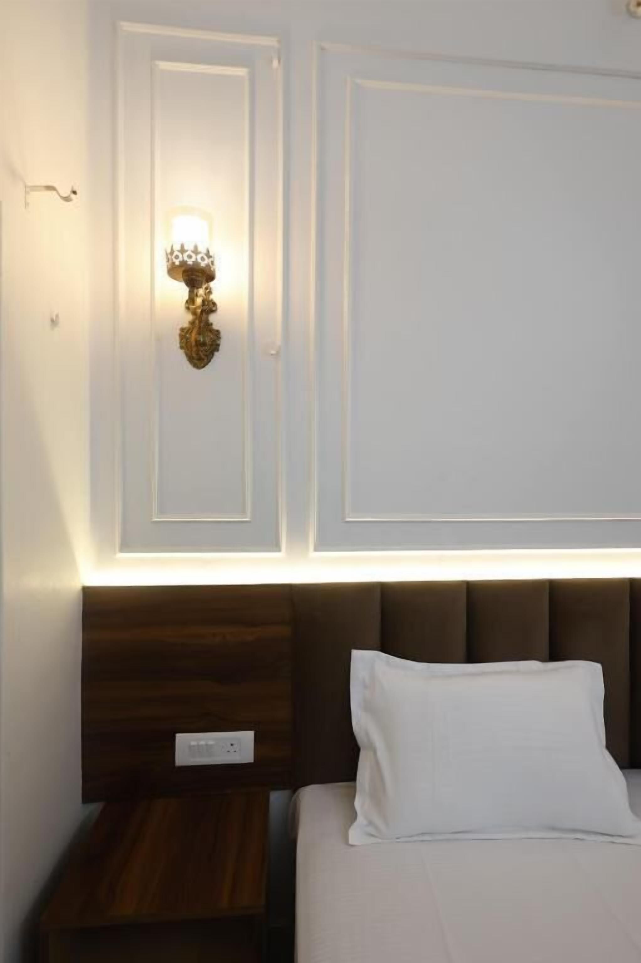 A hotel room with a bed, a nightstand, a wall-mounted light fixture, and a plain wall.