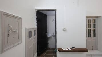 Um corredor com uma porta que dá para um banheiro, uma pequena prateleira de madeira com produtos de higiene pessoal e uma parede branca com um interruptor de luz.