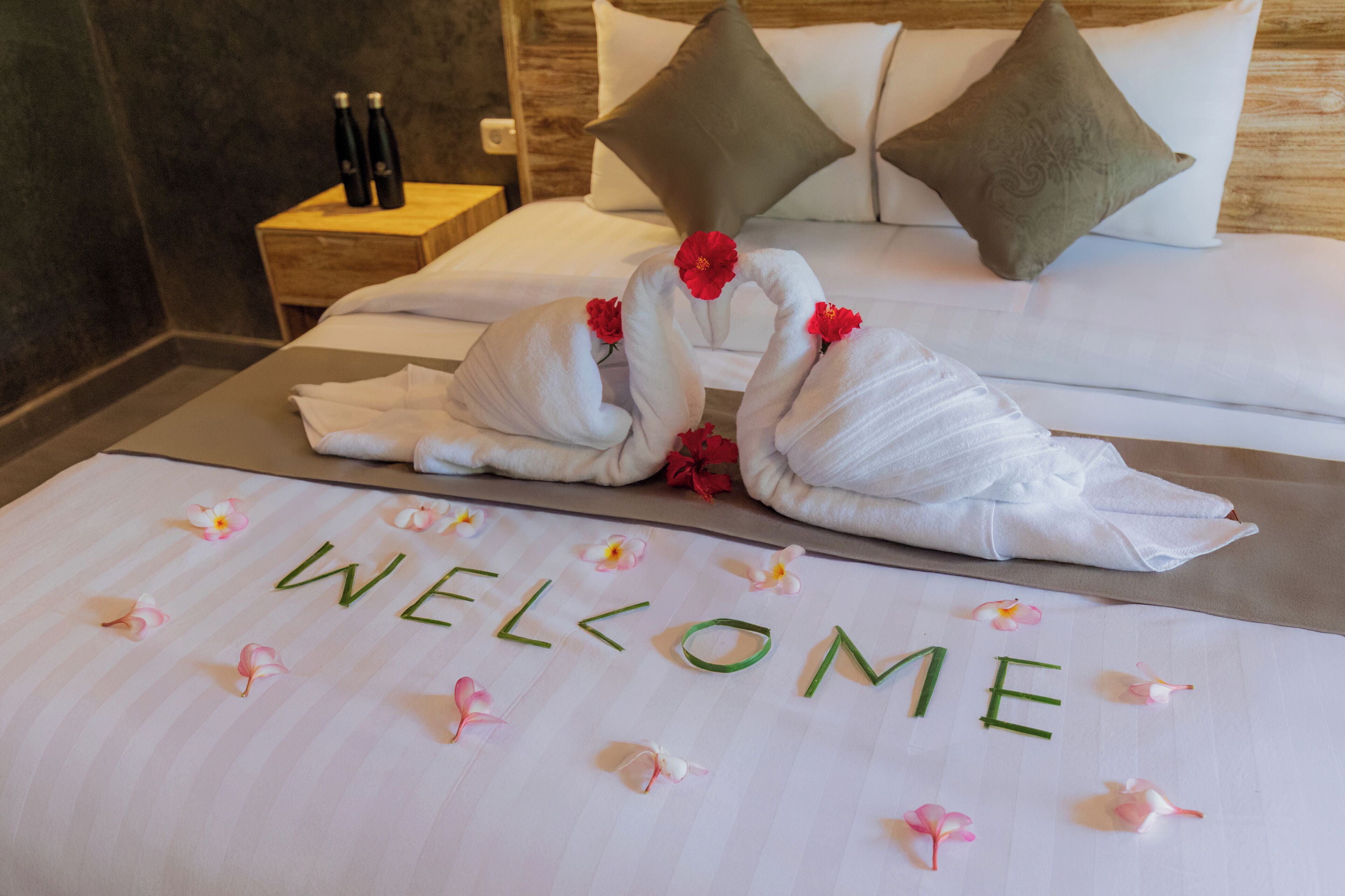 A neatly made bed with towels arranged in swans, a welcome mat with flowers, and a nightstand with bottles.