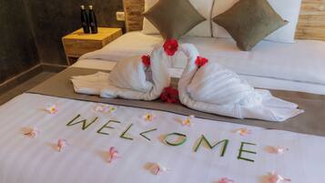 A neatly made bed with towels arranged in swans, a welcome mat with flowers, and a nightstand with bottles.