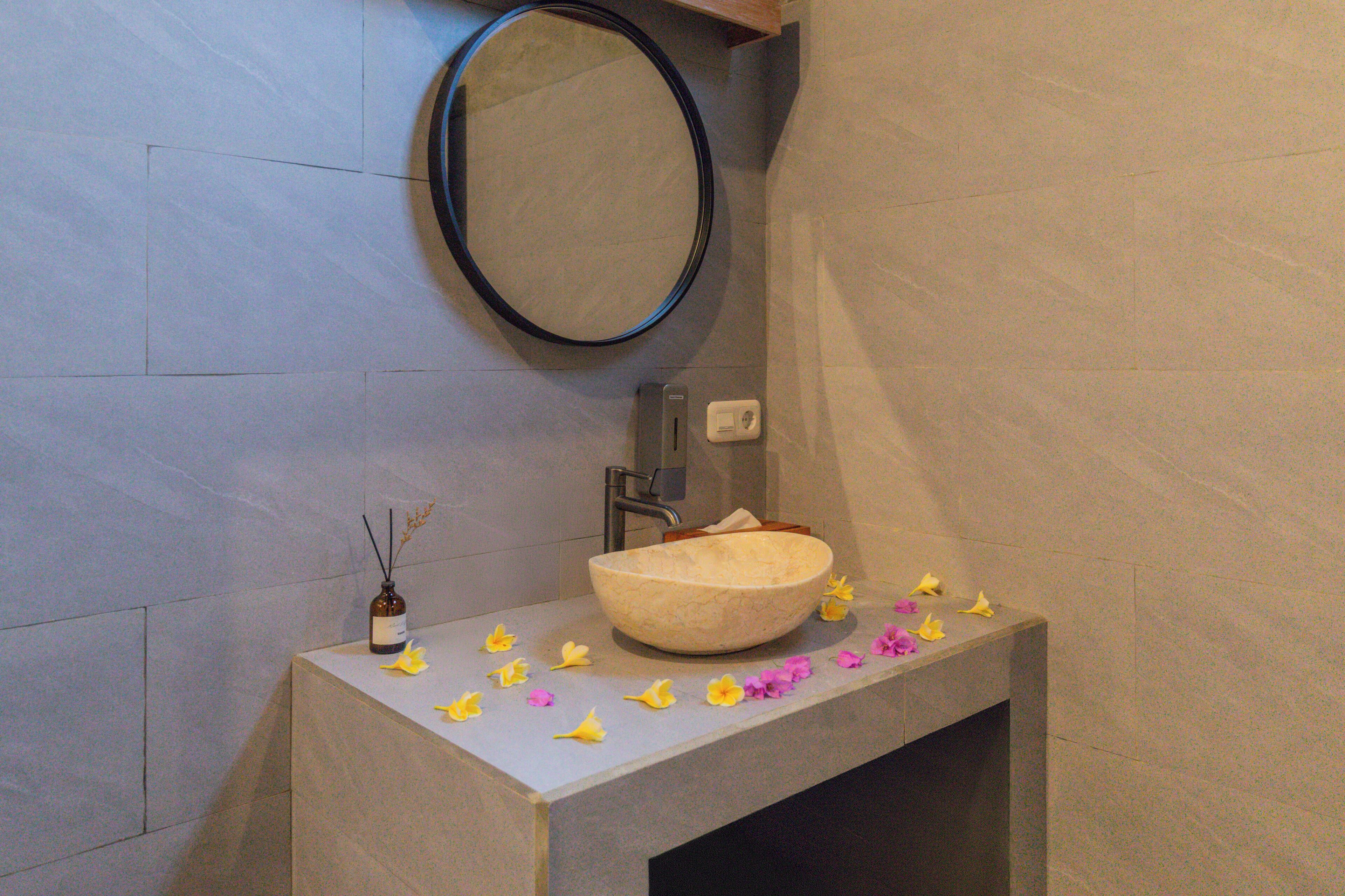 Un baño con un espejo ovalado grande, un lavabo de piedra color beige y una pequeña botella de difusor de aromas.