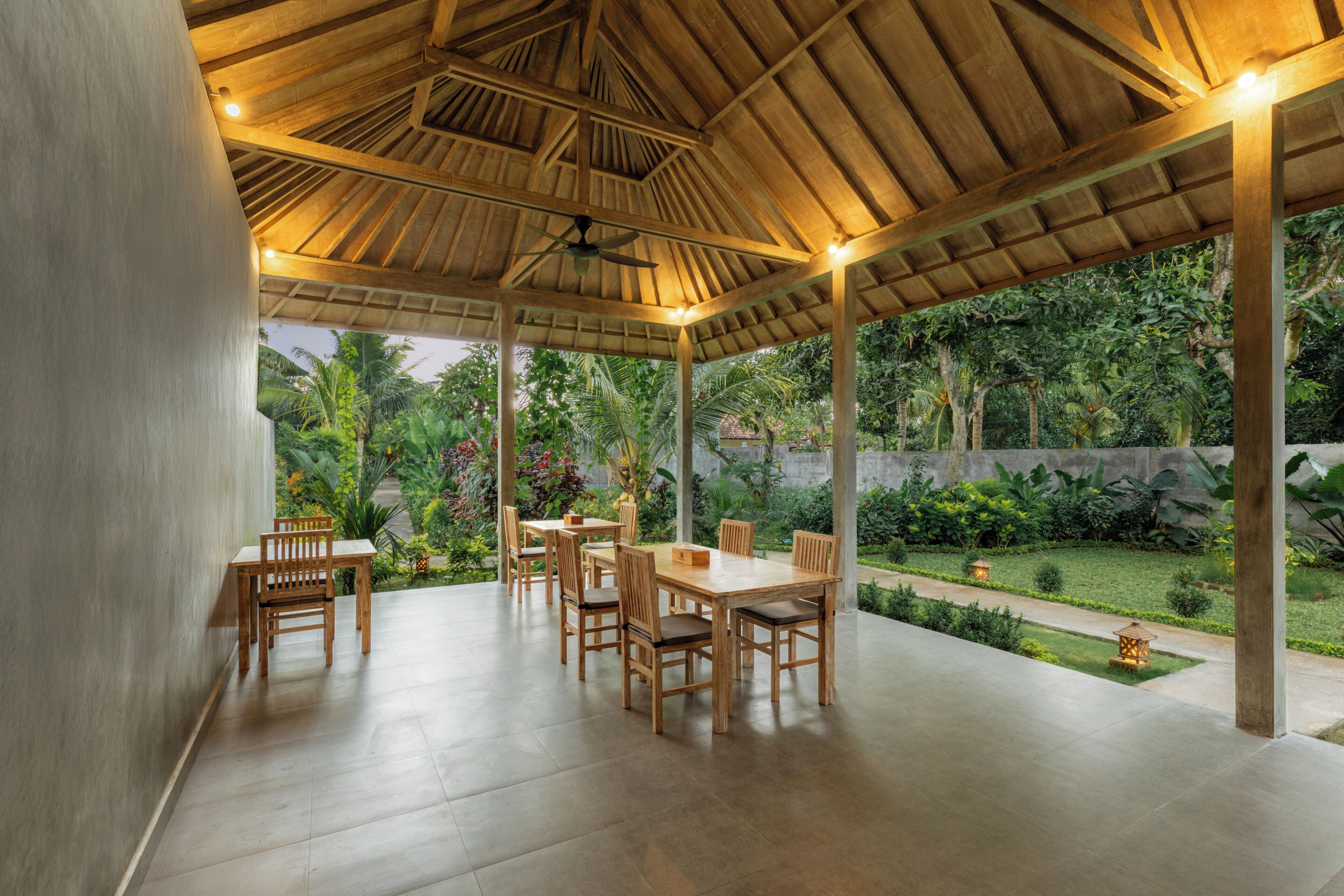 A covered outdoor dining area with wooden tables and chairs, a ceiling fan, and a view of a lush garden.