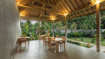 A covered outdoor dining area with wooden tables and chairs, a ceiling fan, and a view of a lush garden.