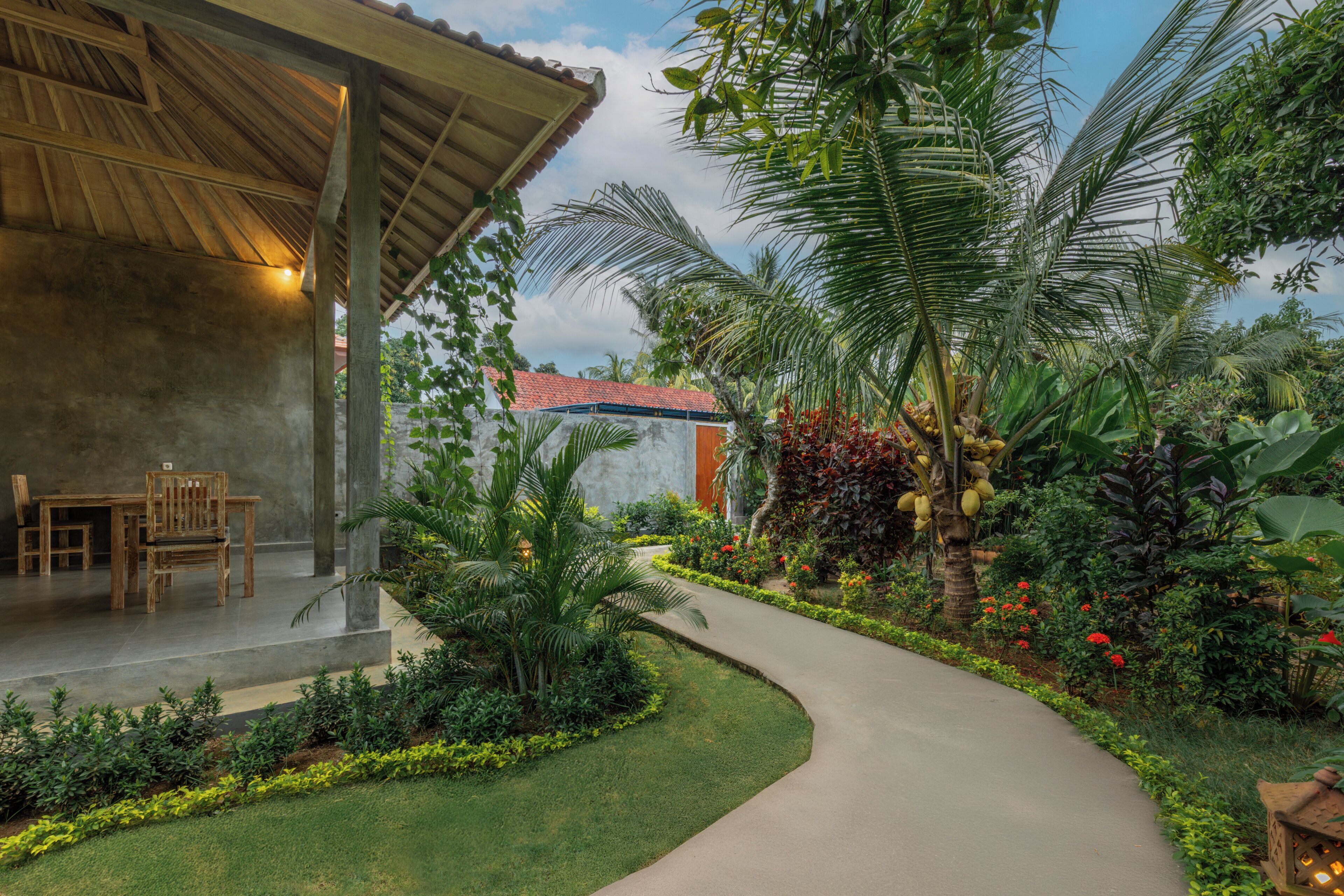 Un espacio exterior cubierto con piso de concreto, una mesa y sillas de madera, y un sendero curvo a través de un jardín con diversas plantas y una palmera.