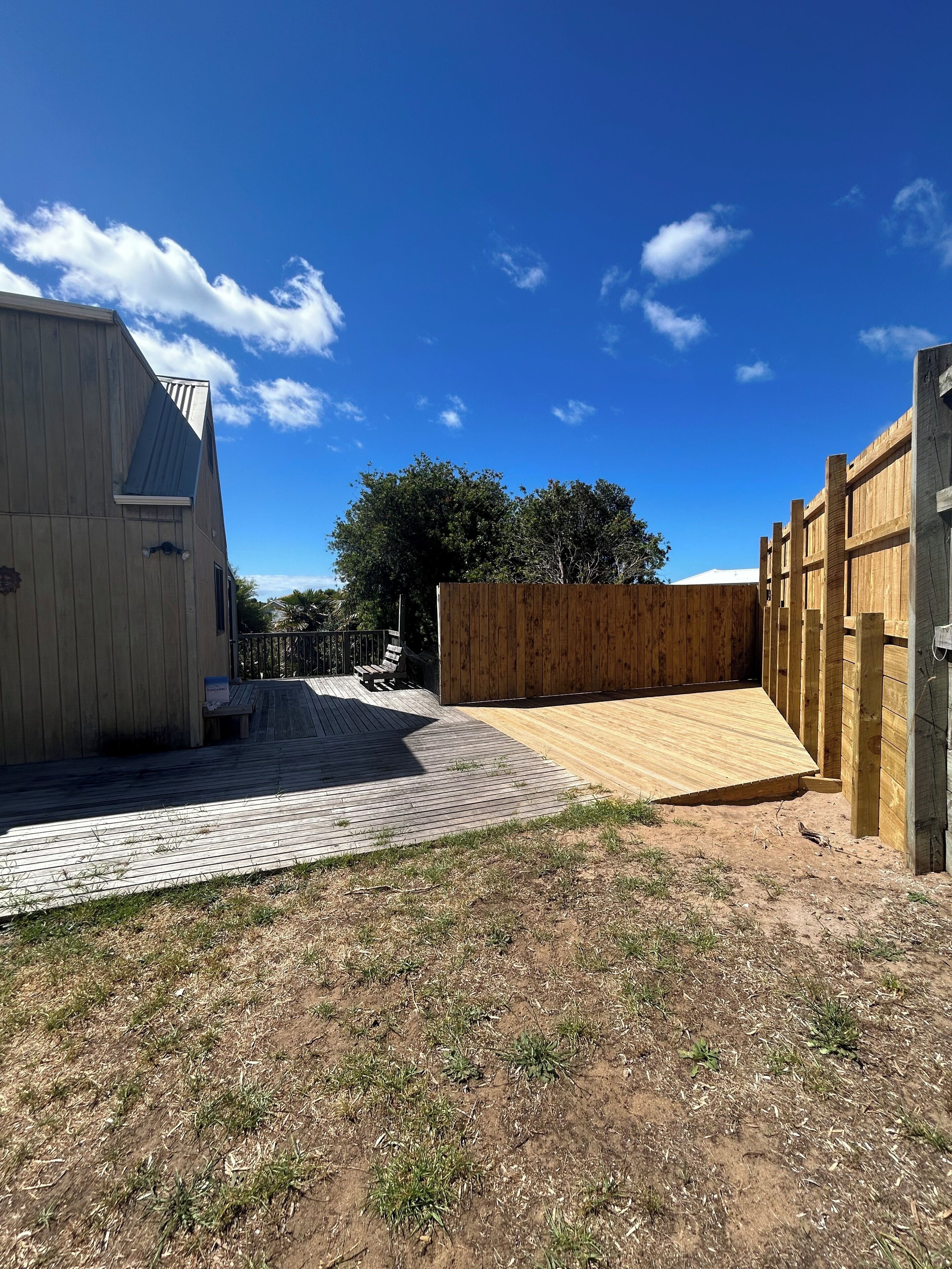 Une terrasse en bois avec une rampe menant à une zone délimitée par une clôture, le tout entouré d’un bâtiment doté d’un toit en pente.