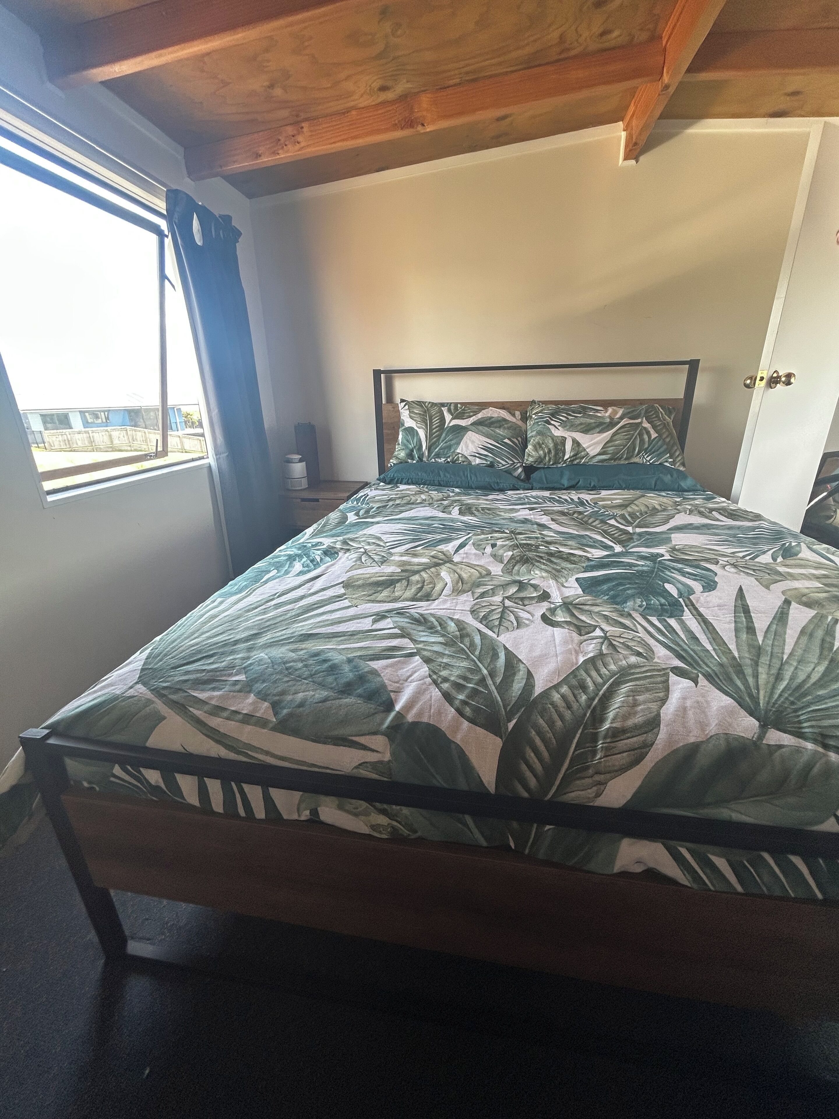 Un lit avec une couvre-lit orné d’un motif de feuilles tropicales, une tête de lit en bois et une fenêtre avec des rideaux.