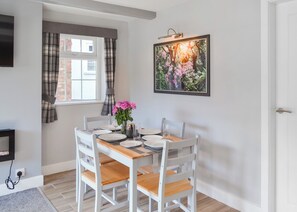 En spiseplads med et bord dækket til fire personer, et vindue med gardiner, et billede på væggen og et fjernsyn på væggen.