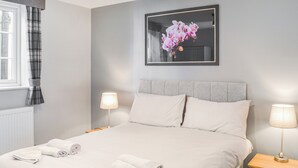 Een net opgemaakt bed met witte lakens en handdoeken, twee nachtlampjes, een bloemistisch kunstwerk in een lijst aan de muur en een raam met ruitgordijnen.