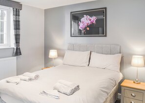 En pænt redt seng med hvide sengetæppe og håndklæder, to sengeborde med lamper, et blomsterbillede i ramme på væggen, og et vindue med ternede gardiner.