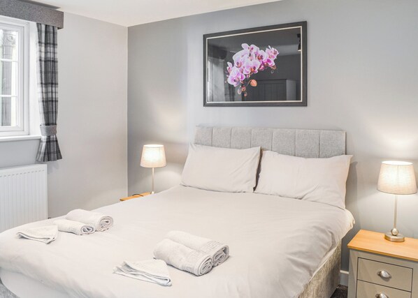 A neatly made bed with white linens and towels, two bedside lamps, a framed floral artwork on the wall, and a window with plaid curtains.