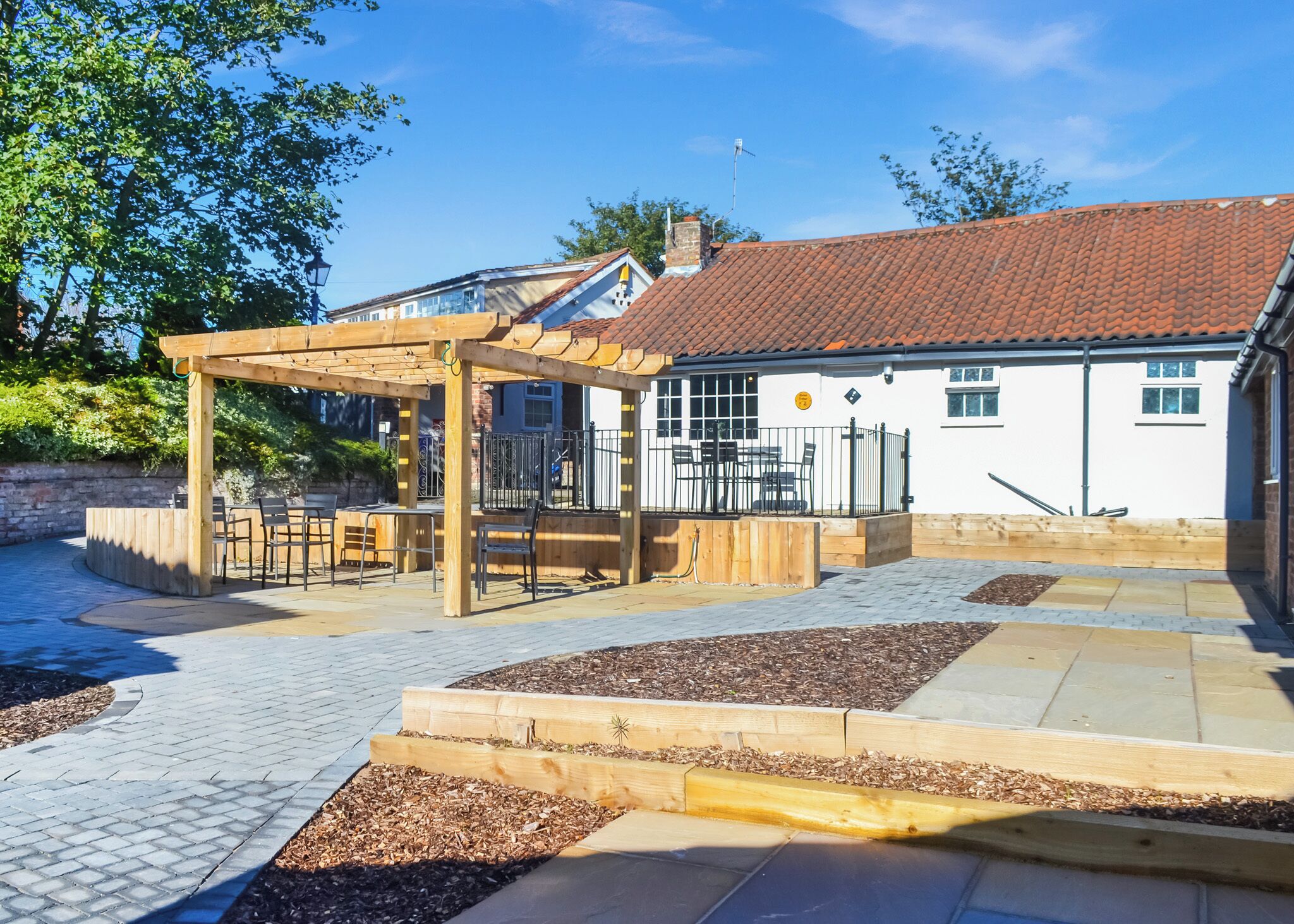 A covered outdoor seating area with a pergola, a fenced play area, and a building with a red-tiled roof.