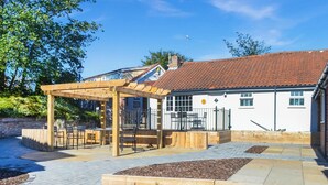 Een overdekt terras met een pergola, een omheind speelgedeelte en een gebouw met een rode pannendak.