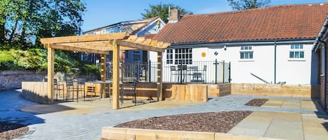 A covered outdoor seating area with a pergola, a fenced play area, and a building with a red-tiled roof.