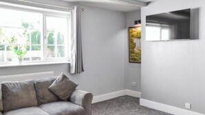 A room with a grey sofa, a radiator, a window with curtains, and a wall-mounted TV. - A beautifully restored single-storey two bedroom cottage sleeping four people. (Filey)