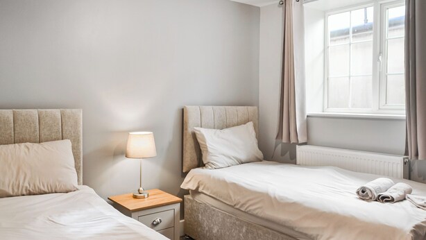 A hotel room with two beds, a nightstand with a lamp, a window with curtains, and a radiator. - A beautifully restored single-storey two bedroom cottage sleeping four people. (Filey)