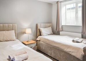 A hotel room with two beds, a nightstand with a lamp, a window with curtains, and a radiator. - A beautifully restored single-storey two bedroom cottage sleeping four people. (Filey)