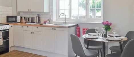Une cuisine moderne avec des armoires blanches, un plan de travail en bois, un four encastré, un four à micro-ondes et un évier. On y trouve également un coin repas avec une table dressée pour deux personnes, des chaises et un vase rempli de fleurs.