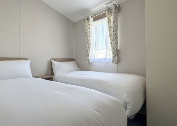 A hotel room with two single beds, a window with patterned curtains, and a plain wall.