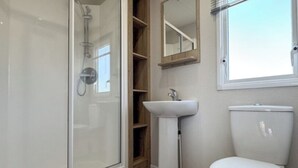 Et badeværelse med et brusebad bag glas, en pedestalvask, et toilet og en hylde i træ.