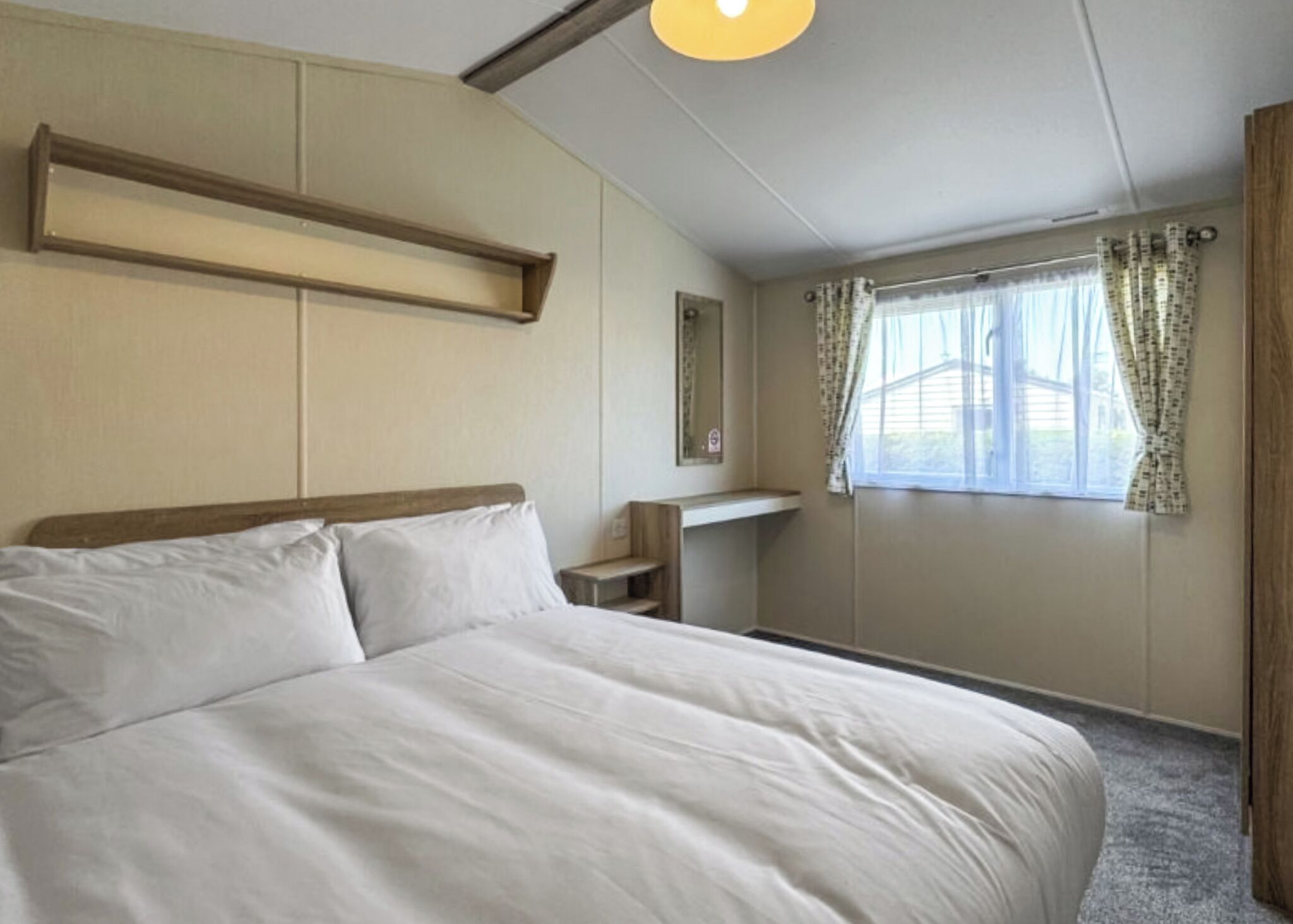 A bedroom with a large bed, a bedside table, a mirror, a window with curtains, and a wooden shelf.