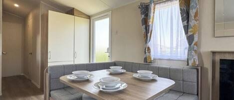 A compact dining area with a wooden table set for four, grey cushioned seating, a window with floral curtains, and a fireplace.