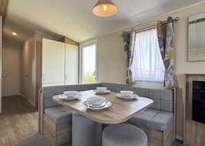 A compact dining area with a wooden table set for four, grey cushioned seating, a window with floral curtains, and a fireplace.