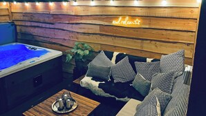 Een gezellig chaletinterieur met een houten wand, een bubbelbad, een bank met gekleurde kussens en een kleine tafel met kaarsen.