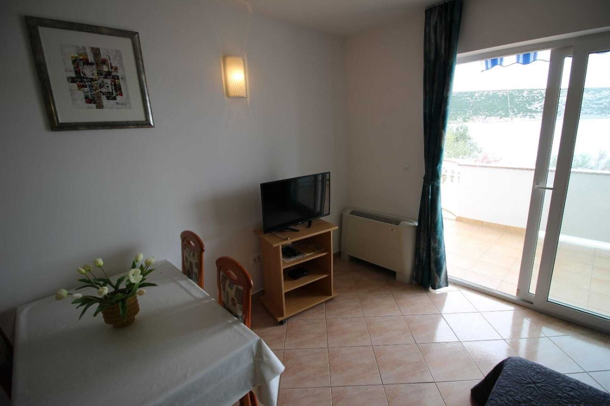 Quarto com uma mesa de jantar, cadeiras, uma TV em um suporte de madeira, uma luminária fixada na parede e uma porta de vidro deslizante para uma varanda.