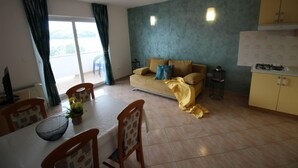 Ein Wohnzimmer mit einer Couch, einem Esstisch, Stühlen, einer kleinen Küchenzeile und Blick durch eine Glastür.