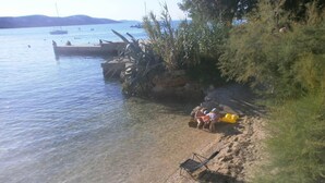 Une plage avec une eau claire et peu profonde, quelques bateaux visibles au loin, et une zone sablonneuse avec une chaise et quelques plantes.
