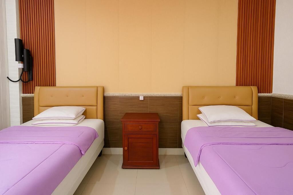 A hotel room with two beds, each with a headboard and a purple bedspread. There is a wooden nightstand between the beds.