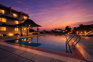 Uma piscina com escada de metal, rodeada por espreguiçadeiras e uma área de estar coberta.