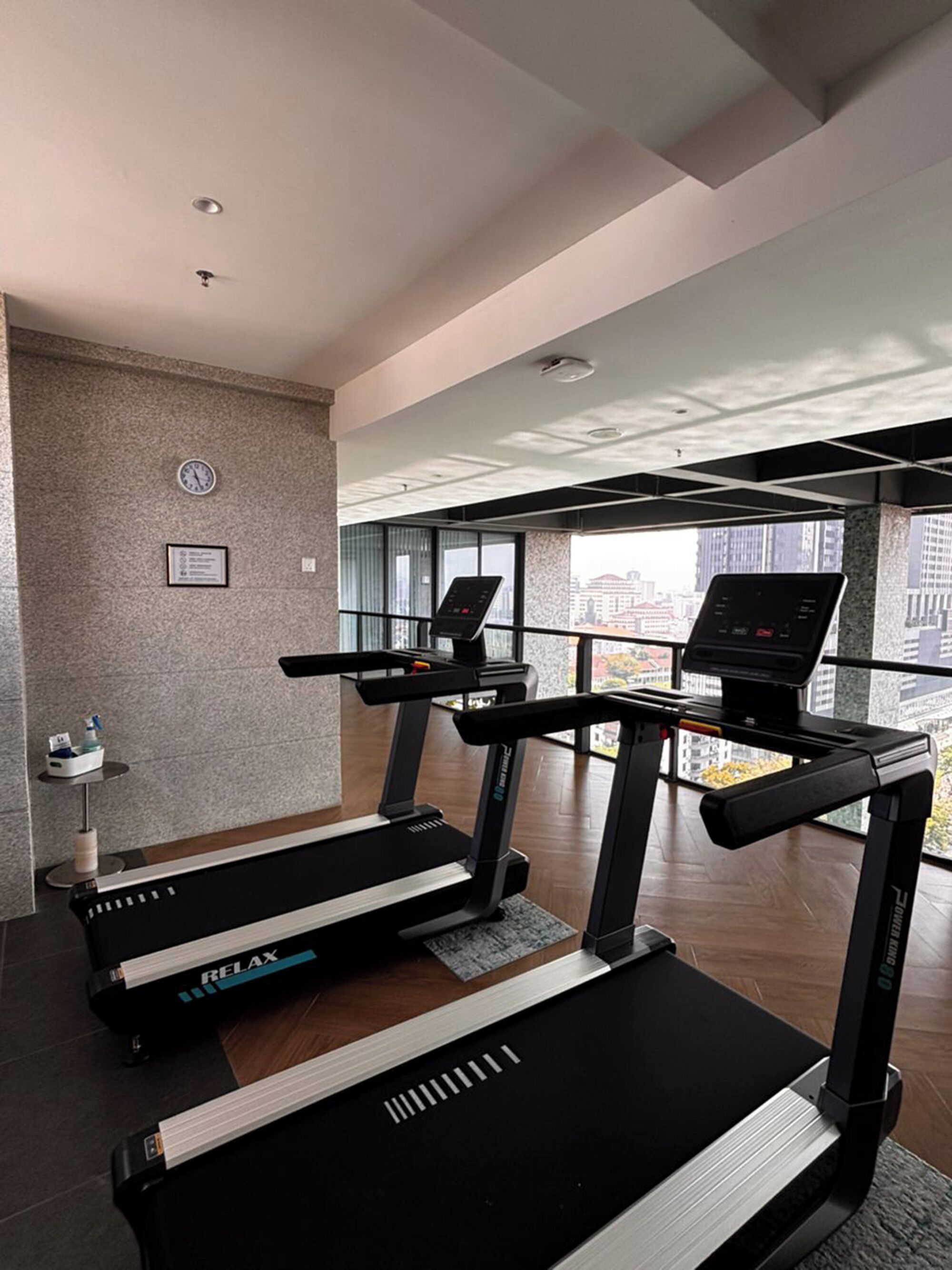 Un gimnasio moderno con cintas de correr, una pantalla digital y vistas a la ciudad.