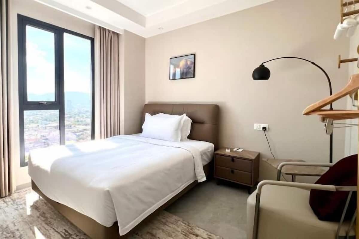 A hotel room with a large bed, a bedside table, a chair, a desk, a lamp, and a view of the city through the window.