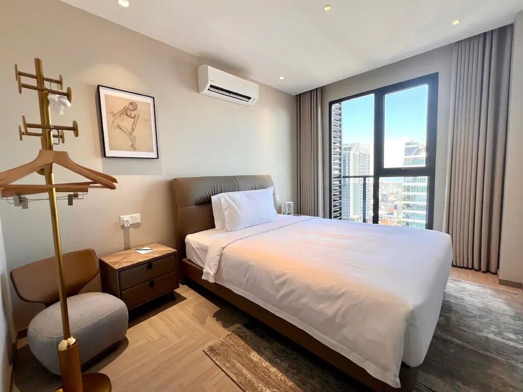 A hotel room with a large bed, a bedside table, a wardrobe, an air conditioning unit, and a window with a city view.