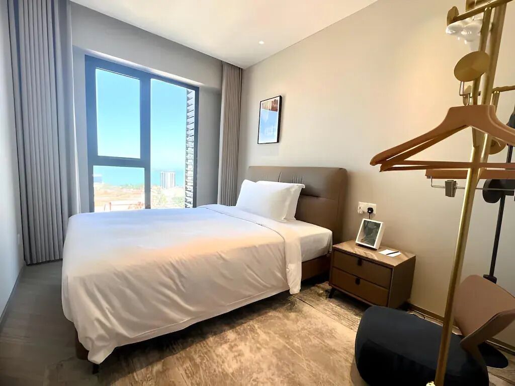 A hotel room with a large bed, a bedside table with a lamp, a chair, and a view of the outdoors.