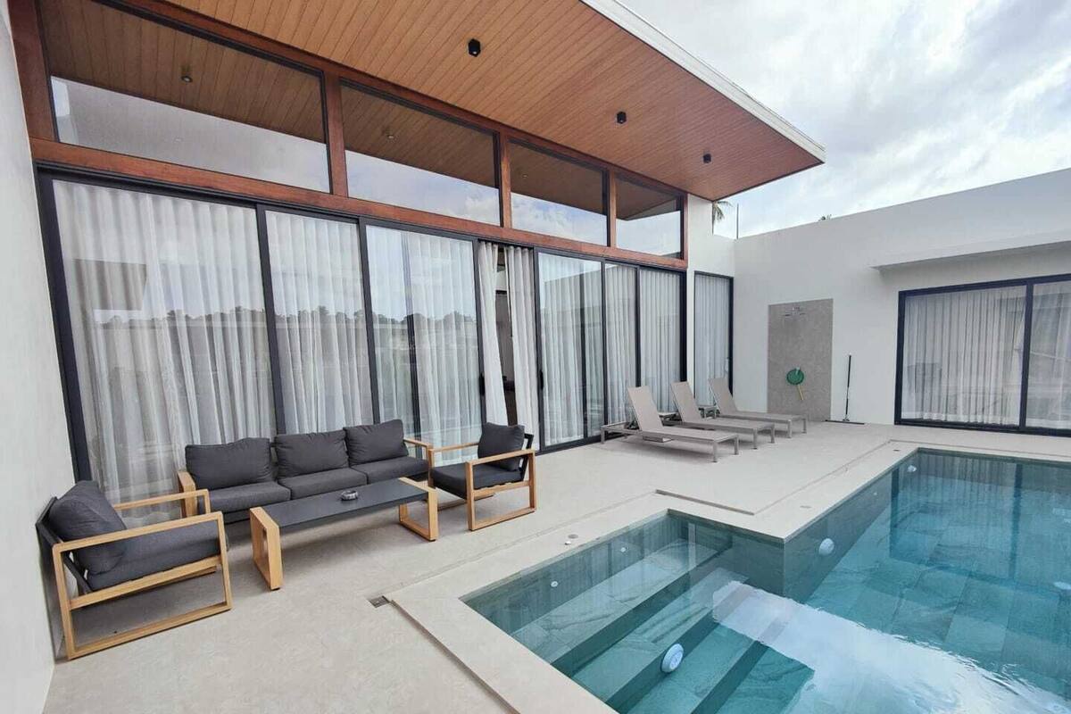 A modern indoor pool area with a glass-enclosed pool, a sofa set, and large windows with sheer curtains.