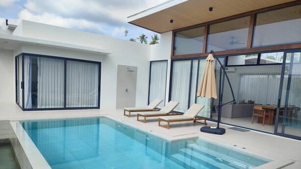 A modern swimming pool with lounge chairs and a sunbed, surrounded by large windows and a glass door. - Samui Luxury villas-BySamui Rental  (Bueang Sam Phan)