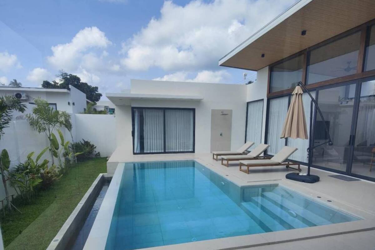 A modern house with a rectangular swimming pool, a covered patio area with two lounge chairs, and a large glass door leading to a covered outdoor space.