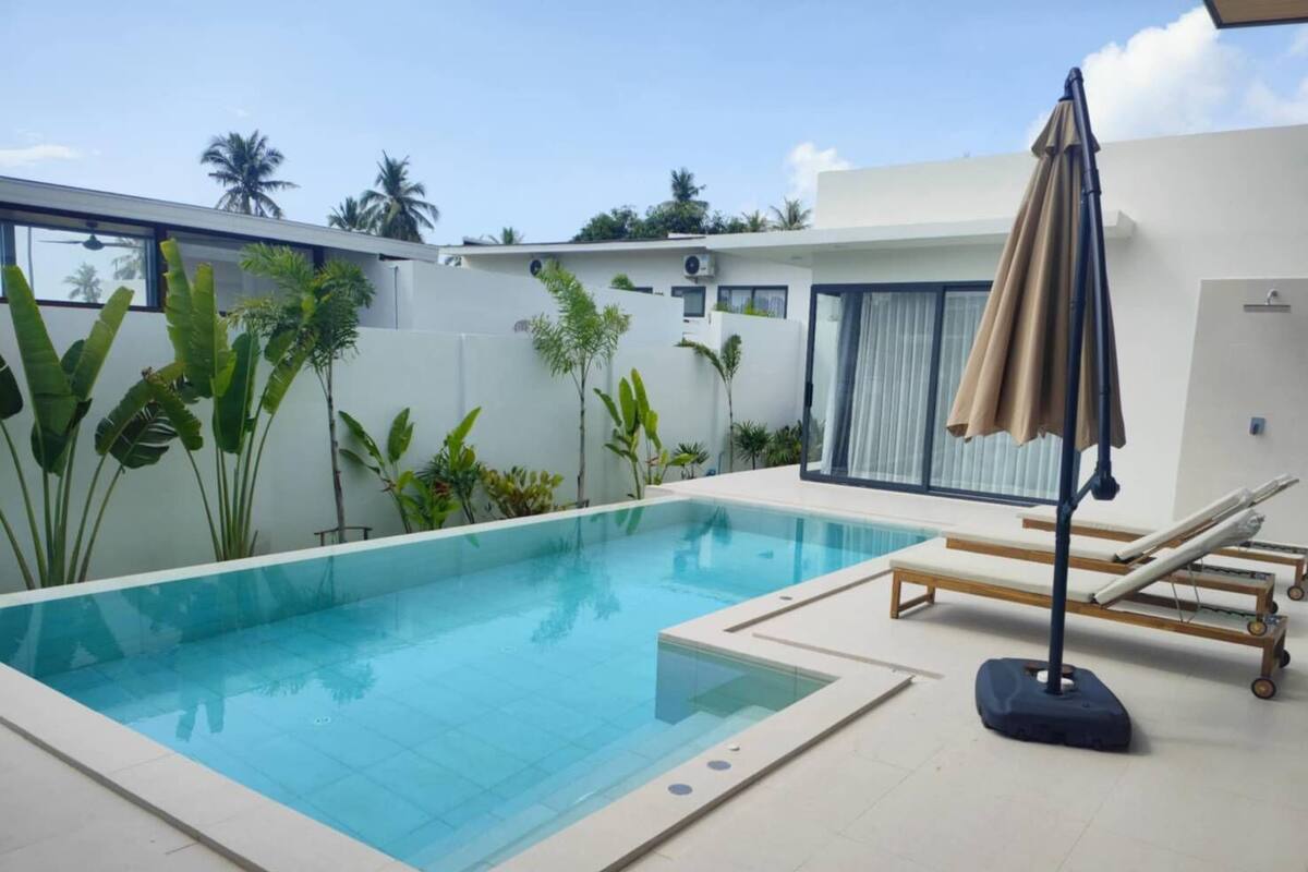 A modern outdoor pool area with a clear blue pool, lounge chairs, and a covered umbrella.