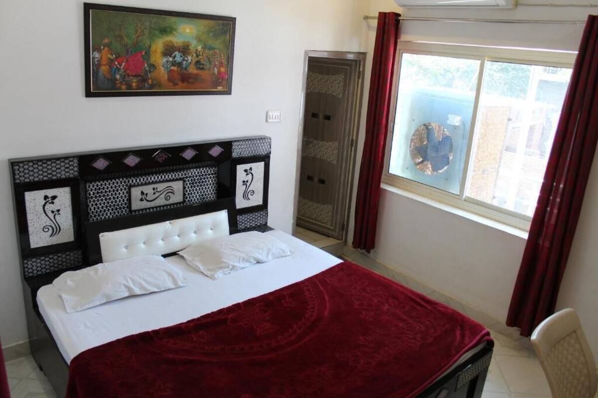 Ein Bett mit roter Decke, ein weißer Kopfseitenverkleidung mit Verzierungen, ein Bild über dem Bett und ein Fenster mit Vorhängen.