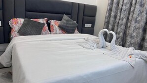 Cama bem arrumada com arranjo de toalhas em formato de coração, cabeceira escura, almofadas estampadas e cortina cinza.