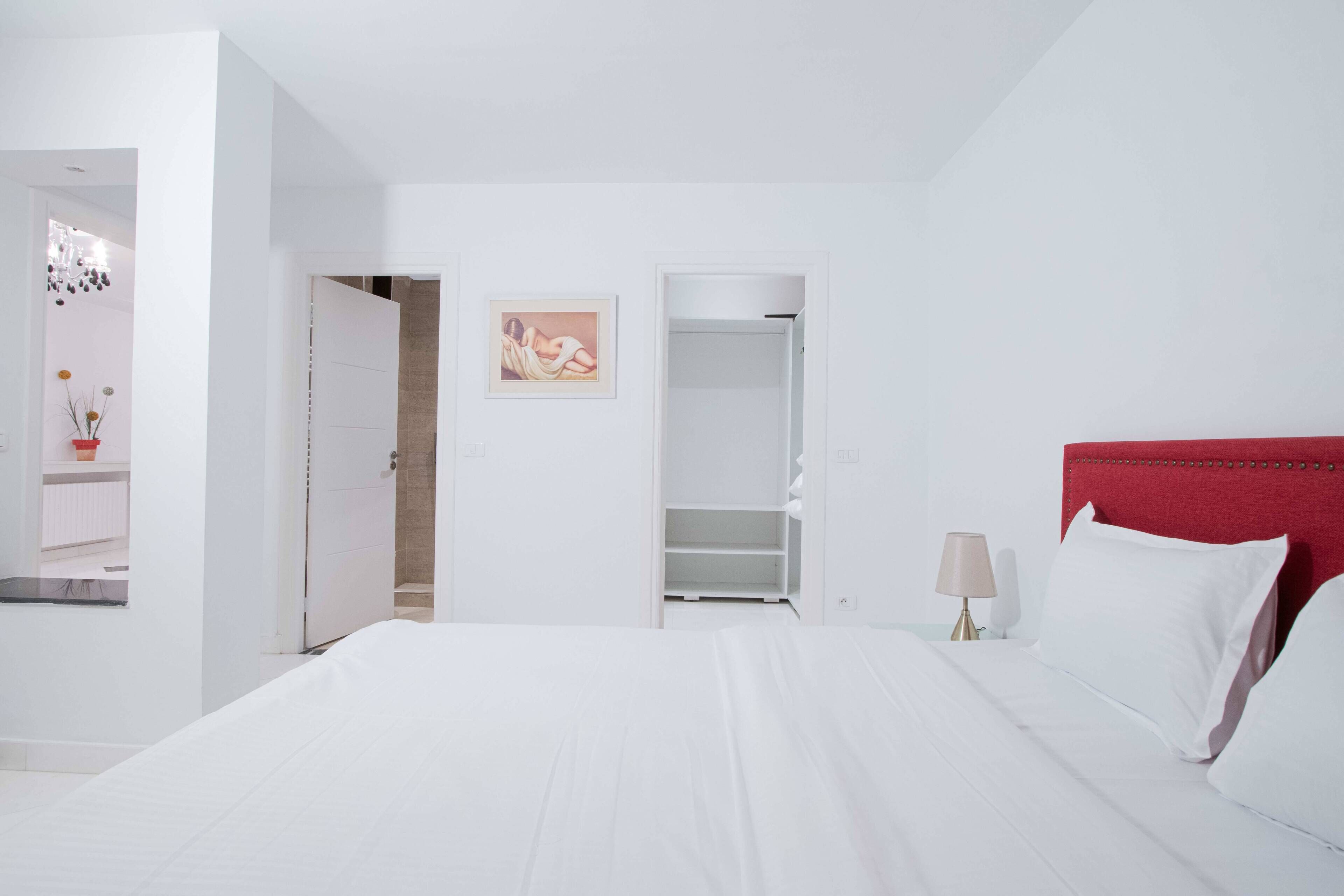 Habitación de hotel con una cama grande, cabecera roja, mesita de noche con lámpara y un armario.