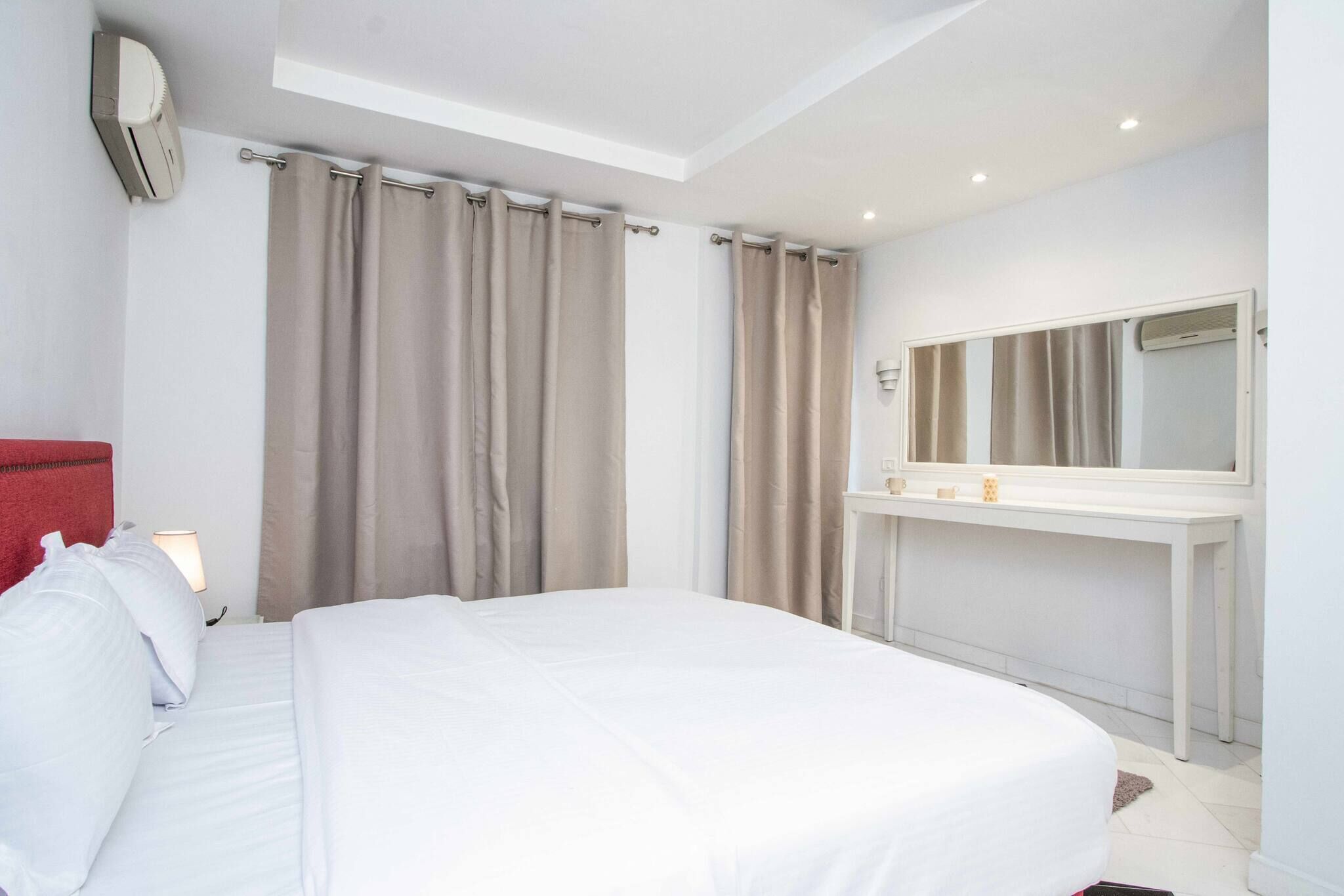 A hotel room with a large bed, a bedside table with a lamp, a mirror above the table, and beige curtains.
