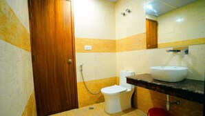 Ein Badezimmer mit Holztür, Toilette in Weiß, Waschbecken mit Spiegel darüber und einem roten Eimer auf dem Boden.