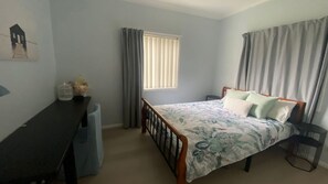 A bedroom with a bed, a desk, a window with blinds, and a picture on the wall.