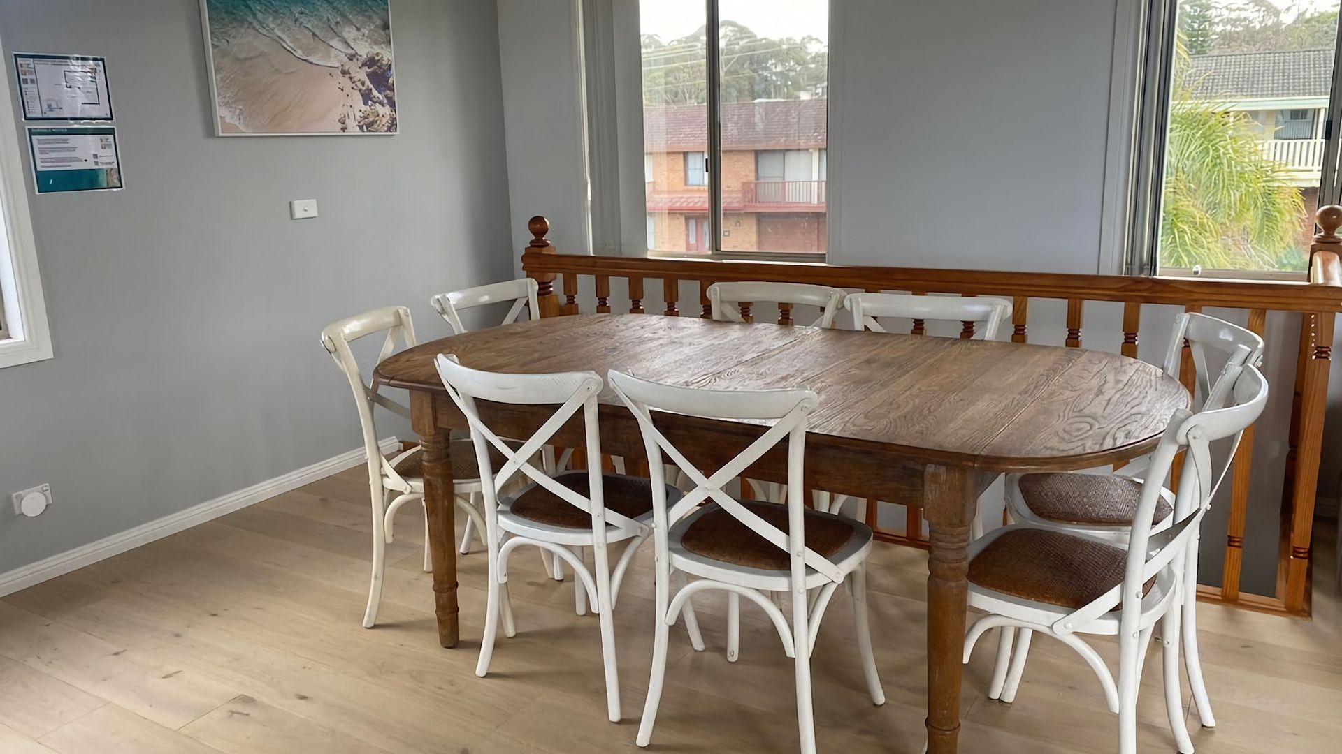 A dining area with a wooden table and white chairs, a window with a view of buildings, and a painting on the wall.