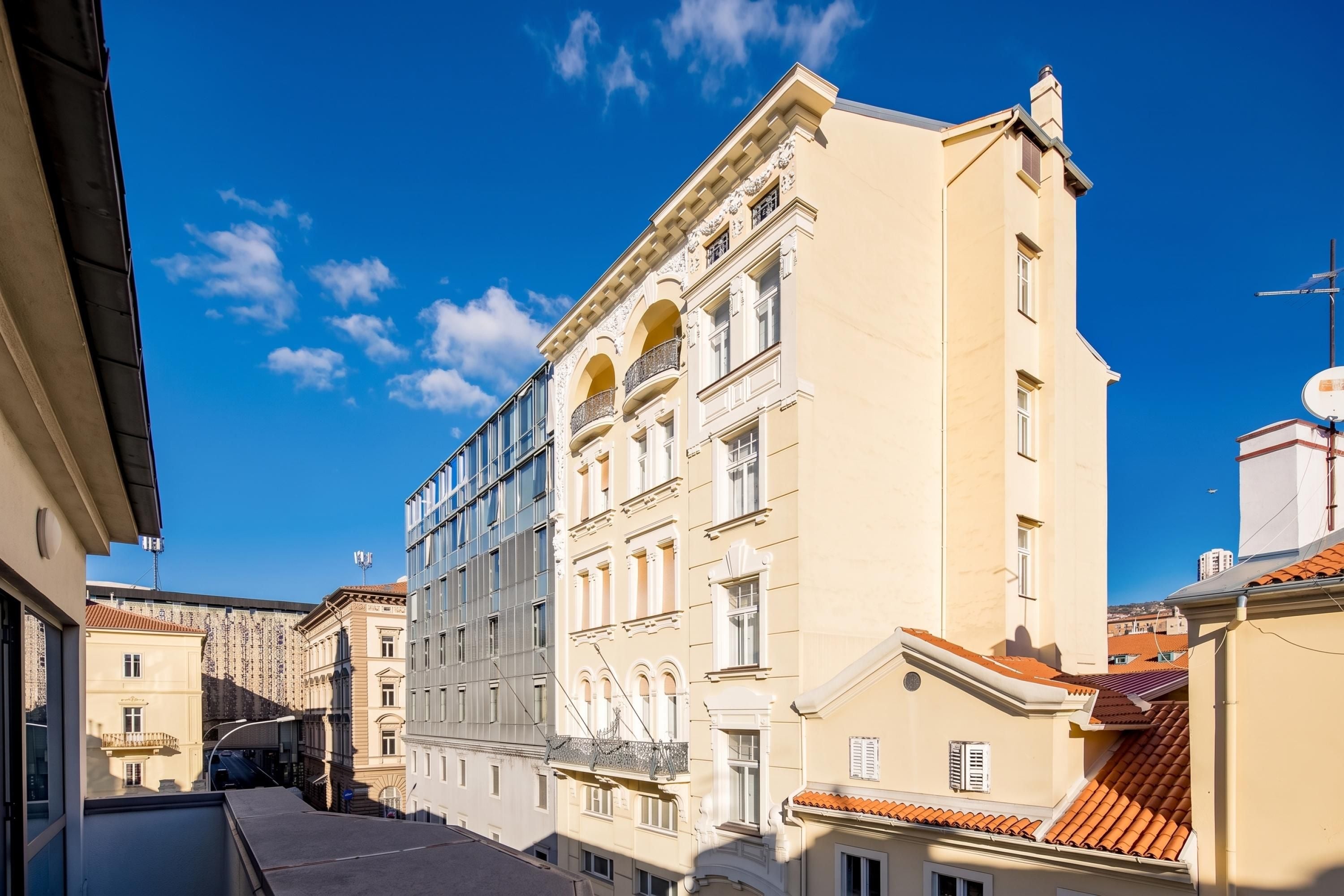 A narrow street with buildings featuring classical architecture, including arched windows and decorative moldings.