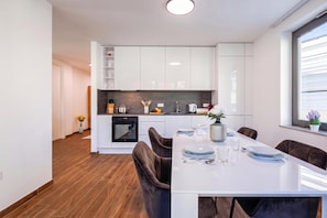 A modern kitchen with white cabinets, a built-in oven, and a dining area with a white table and brown chairs. - City Apartments (Rijeka)