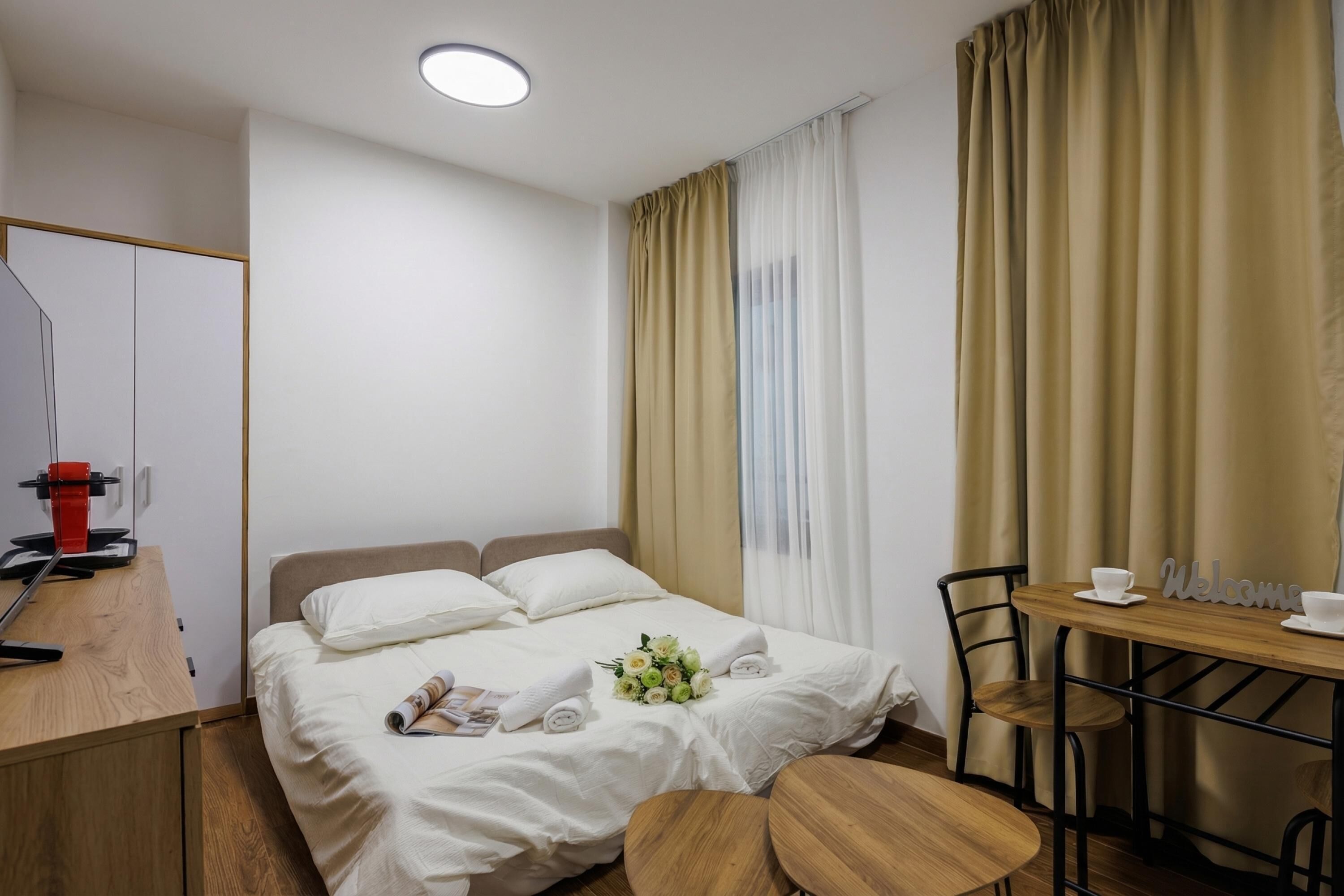 A hotel room with a bed, a desk with a computer, a chair, a table with a 'Welcome' sign, and a window with curtains.